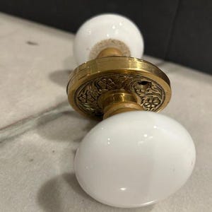 May include: Antique door knob with white porcelain egg-shaped handles and a brass rosette. The brass rosette has an ornate floral design. The door knob is a vintage style.