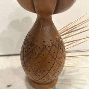 May include: A brown wooden toothpick holder shaped like a pineapple with a flower-shaped top. The body features a cross-hatched diamond pattern with small holes for inserting wooden toothpicks, several of which are currently placed in the side.