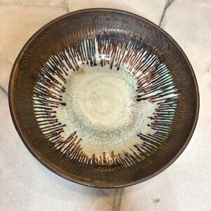 May include: A round ceramic bowl with a textured brown exterior and a light green and white interior. The interior features a pattern of dark brown lines radiating from the center, creating a unique design.