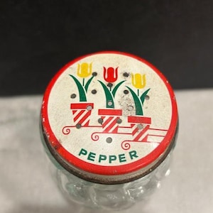 May include: A vintage pepper shaker with a round, red-rimmed lid. The lid features a white background with three potted tulips in yellow and red. The word "PEPPER" is printed in green. The clear glass body is visible.