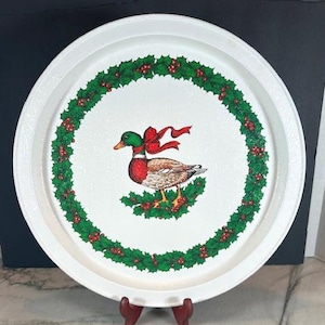 May include: White, round serving tray featuring a duck design within a holly wreath. The duck has a green head, red chest, and brown body, with a red bow. The wreath is green with red berries.