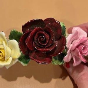 Może przedstawiać: Ceramiczna kompozycja kwiatowa z trzema różami w różnych kolorach. Kompozycja zawiera ciemnoczerwoną różę, jasno-różową różę i żółtą różę, wszystkie z zielonymi liśćmi. Róże są prawdopodobnie częścią przedmiotu dekoracyjnego.