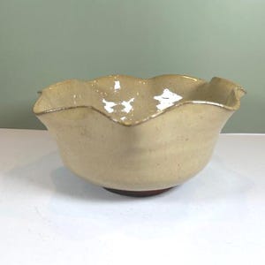 May include: A handmade ceramic bowl with a ruffled rim. The bowl is a light beige color with a darker brown base. The rim has a slightly darker, golden-brown edge. The bowl is likely used for serving food or as a decorative piece.