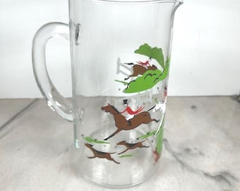 May include: Clear glass pitcher with a handle and a spout. The pitcher features a colorful illustration of a fox hunt scene with horses, riders, and hounds. The design includes green foliage and a white fence.