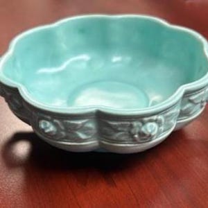 May include: A light blue ceramic bowl with a scalloped rim and two handles. The bowl has a floral design around the base and is sitting on a wooden surface. This decorative bowl is suitable for serving.