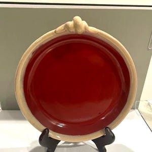 May include: A round, red ceramic plate with a cream-colored rim and decorative accents. The plate is displayed on a black stand, showcasing its design. The plate's center is a deep red, contrasting with the lighter rim.