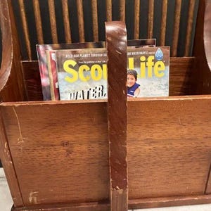 May include: A brown wooden magazine rack featuring a central vertical handle and a slatted back design. The rack contains two magazines, one of which displays the text 'Scout Life' and 'HELP OUR PLANET THROUGH OUR WATERWAYS' on its front cover.