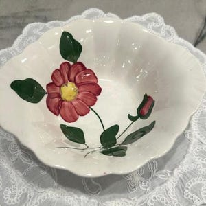 May include: A white ceramic bowl with a scalloped edge featuring a hand-painted design of a large red flower with a yellow center, a small red bud, and several green leaves. The bowl has a glossy finish and a smooth, curved interior surface.