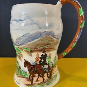 May include: Cream-colored ceramic mug with a landscape scene and a horse-drawn carriage. The handle is brown with green and purple accents. The mug features a painted scene of a horse and carriage on a road with mountains in the background.
