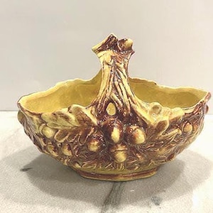 May include: A ceramic basket with a handle, featuring a textured design of acorns and leaves. The basket is primarily yellow with brown accents, creating a rustic and natural aesthetic. The handle is shaped like a tree trunk.