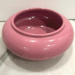 May include: A smooth, glossy, rose-pink ceramic bowl with a rounded, bulbous body and a wide, circular opening. The interior of the bowl is finished in the same solid pink color, featuring a clean, minimalist design with a curved rim.