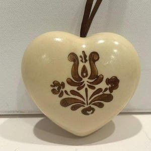May include: A cream-colored ceramic heart ornament featuring a brown painted floral motif on the front. The heart has a smooth, glossy finish and is suspended by a thin brown ribbon attached to the top center of the piece.