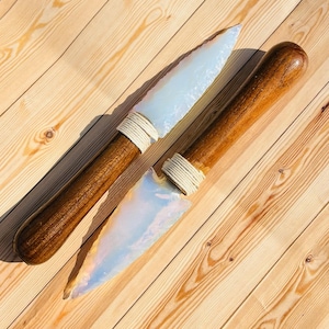 Puede incluir: Dos cuchillos hechos a mano con mangos de madera y hojas opalescentes. Los mangos de color marrón oscuro están envueltos con un cordón blanco cerca de la hoja. Las hojas tienen una apariencia nacarada y translúcida.