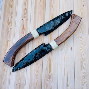 Peut inclure: Deux couteaux en obsidienne avec des manches en bois. Les lames sont noires et les manches sont brun foncé avec un design incurvé. Les manches sont enveloppés d'une corde blanche près de la lame. Les couteaux sont présentés sur une surface en bois brun clair.
