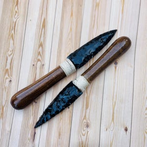 May include: Two obsidian spearheads with dark wooden handles, wrapped with natural twine. The blades are a deep black, contrasting with the polished brown handles. The items are displayed on a light wood grain surface.