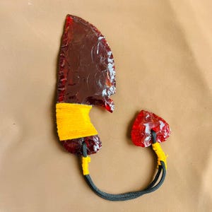 Puede incluir: Un cuchillo artesanal de vidrio rojo con un mango curvo envuelto en hilo amarillo. Un trozo de vidrio rojo más pequeño, en forma de corazón, está conectado por un cordón negro. El cuchillo y el corazón se exhiben sobre una superficie marrón.