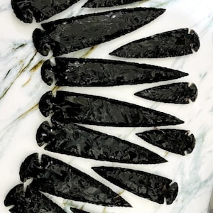 以下が含まれることがあります： 白とグレーの大理石の表面に配置された、さまざまなサイズの黒曜石の矢じりのコレクション。矢じりは光沢のある反射仕上げで、鋭いエッジと詳細な職人技を示しています。コレクションにはいくつかのサイズが含まれています。