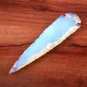 Puede incluir: Una punta de flecha translúcida de color azul claro con una punta afilada y bordes irregulares. La punta de flecha está hecha de un material vítreo y se encuentra sobre una superficie de madera marrón. Un artículo artesanal único.