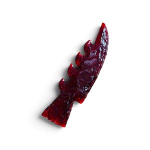 May include: A deep red, translucent arrowhead with a serrated edge and a pointed tip. The surface features a chipped, flaked texture characteristic of knapped stone, creating a glossy, glass-like appearance with varying shades of dark crimson.