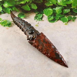 May include: A handcrafted dagger with a reddish-brown blade and a dark brown handle. The handle features metal bands and a black accent. The blade has a textured appearance, and the overall design suggests a historical or fantasy theme.