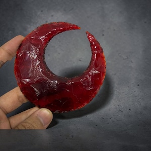 Puede incluir: Un objeto con forma de media luna tallado en piedra roja profunda y translúcida con un acabado pulido y brillante. La superficie muestra texturas naturales de escamas irregulares y bordes afilados y definidos en toda la pieza.