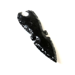 May include: A black obsidian arrowhead with a notched design. The arrowhead has a sharp, pointed tip and a smooth, polished surface. The edges are finely worked, and the overall shape is symmetrical. The arrowhead is set against a white background.
