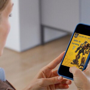 May include: A person holding a blue smartphone displaying a birthday invitation. The invitation features a yellow and black design with a robot illustration and the text "John is turning 7".