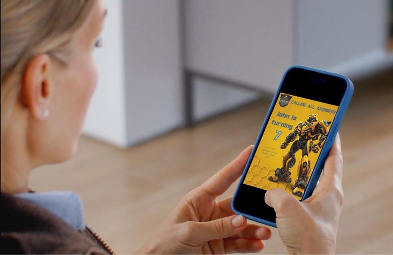 May include: A person holding a blue smartphone displaying a birthday invitation. The invitation features a yellow and black design with a robot illustration and the text "John is turning 7".