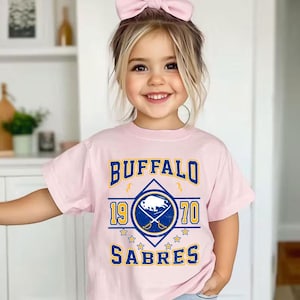 Op de afbeelding: Een lichtroze t-shirt met de tekst "BUFFALO SABRES 1970" in blauw en goud. Het shirt heeft een afbeelding van een buffel en gekruiste zwaarden. Het t-shirt wordt gedragen door een jong kind met een roze strik in het haar.