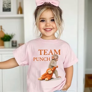 Peut inclure: T-shirt rose clair avec l'inscription "TEAM PUNCH" en marron. Le motif représente un singe portant un singe en peluche. L'enfant porte un nœud rose dans les cheveux.