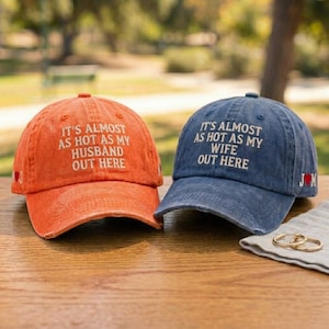 May include: Two baseball caps, one orange and one blue, are displayed. The orange cap features the text "IT'S ALMOST AS HOT AS MY HUSBAND OUT HERE". The blue cap has the text "IT'S ALMOST AS HOT AS MY WIFE OUT HERE" embroidered in white.