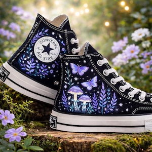 Puede incluir: Zapatillas altas negras con cordones y suelas blancas. Las zapatillas presentan un diseño colorido de mariposas, setas y follaje. El logotipo de Converse All Star es visible en el lateral.