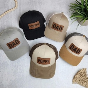 Könnte beinhalten: Fünf Trucker-Caps in verschiedenen Farben, jeweils mit einem Lederpatch mit den Aufschriften "DAD", "POPPY" und "PAWPAW". Die Caps haben Mesh-Rücken und gebogene Schirme, ideal für den Alltag.