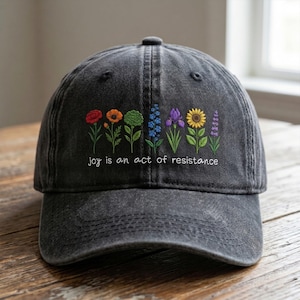 Può includere: Un cappellino da baseball grigio antracite con una fila ricamata di fiori di campo colorati, tra cui tonalità rosse, arancioni, verdi, blu, viola e gialle. Sotto i fiori, il testo "joy is an act of resistance" è ricamato in bianco.