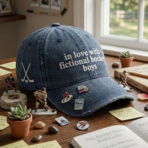 Może przedstawiać: Dżinsowa czapka bejsbolowa z napisem "in love with fictional hockey boys" wyhaftowanym na biało. Czapka ma białe grafiki hokejowych kijów i kilka przypinek o tematyce hokejowej. W pobliżu mały sukulent w doniczce i figurki hokejowe.