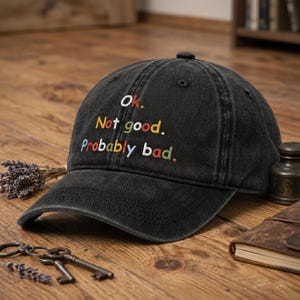 Peut inclure: Casquette de baseball noire avec le texte brodé "Ok. Not good. Probably bad." en blanc, vert, bleu et rouge. La casquette est faite d'un tissu usé et texturé.