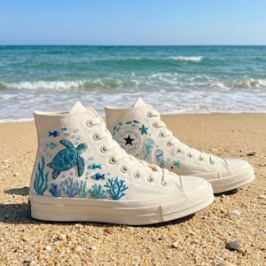 Könnte beinhalten: Cremefarbene High-Top-Sneaker mit maritimen Stickereien. Das Design umfasst eine türkisfarbene Meeresschildkröte, blaue Fische und Korallen. Das Converse All Star-Logo ist sichtbar. Die Schuhe stehen auf einem Sandstrand.