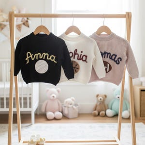 May include: Three children's sweaters hanging on a wooden rack. The sweaters are black, white, and pink. Each sweater has a name embroidered on it, with a sports-themed graphic. The black sweater says "Anna" with a golf ball. The white sweater says "Sophia" with a basketball. The pink sweater says "Thomas" with a football.