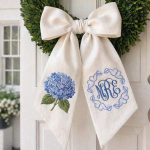 May include: A cream-colored door bow with a large bow and long tails. One tail features a blue hydrangea embroidery, the other a blue monogram within a scalloped border. The bow is set against a green wreath.