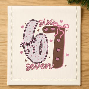 May include: A cream-colored square fabric with the numbers "67" and the word "seven" embroidered in pink. The number six is light pink with sprinkles and a pink bow. The number seven is brown with pink hearts and a pink bow.