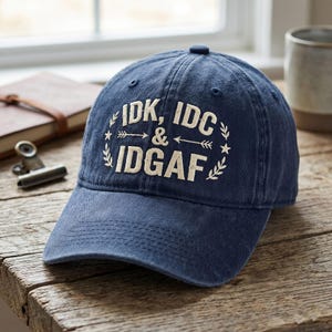 May include: Navy blue baseball cap with the text "IDK, IDC & IDGAF" in off-white lettering. The text is surrounded by a decorative wreath of leaves, stars, and arrows. The cap is made of a soft, worn material.