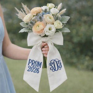 May include: A floral bouquet featuring peach, white, and blue flowers, tied with a white ribbon. The ribbon displays the text "PROM 2026" and a navy blue monogram. The bouquet includes greenery and dried grasses.