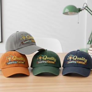 May include: Several baseball caps in shades of grey, orange, green, and navy. Each cap has the embroidered text "Quality Learning Center" with a treehouse design. The caps are arranged on a wooden surface.