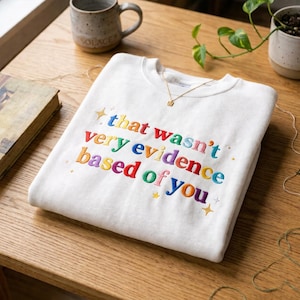 Puede incluir: Sudadera blanca con la frase bordada "that wasn't very evidence based of you" en colores arcoíris. Un collar de oro con un pequeño colgante cuadrado descansa sobre la sudadera. La sudadera está doblada sobre una superficie de madera.