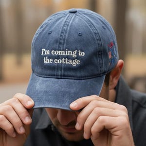 Könnte beinhalten: Eine blaue Baseballkappe mit dem gestickten Text "I'm coming to the cottage" in Weiß. Die Kappe hat einen gebogenen Schirm und ein kleines gesticktes Design an der Seite. Der Hut ist aus einem weichen, abgenutzten Material.