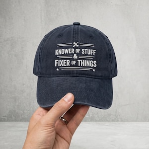 May include: A navy blue baseball cap with the text "KNOWER OF STUFF & FIXER OF THINGS" in white. The text is arranged in two lines with a wrench and screwdriver icon above the top line. The cap has a curved peak.