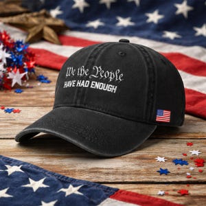 May include: Black baseball cap with the text "We the People HAVE HAD ENOUGH" embroidered in white. The cap features a small American flag patch and is set against a backdrop of the American flag and patriotic decorations.