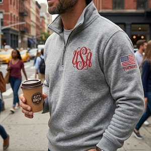 Può includere: Una felpa grigia con zip a un quarto con monogramma rosso e toppa con bandiera americana con la scritta "THOMAS". La felpa ha un collo alto e una cerniera argentata. La persona tiene in mano una tazza di caffè City Brew.