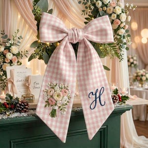 Op de afbeelding: Een grote roze en witte ruitjesstrik met lange linten, geborduurd met een bloemenboeket en een marineblauwe letter "H". De strik maakt deel uit van een bruiloftsdecoratie, met bloemen en kaarsen op de achtergrond.