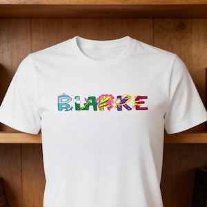 Peut inclure: T-shirt blanc avec le nom "BLARKE" en lettres colorées et à motifs sur la poitrine. Les lettres sont dans des tons de bleu, vert, jaune, rose et violet. Le t-shirt est présenté sur une étagère en bois.